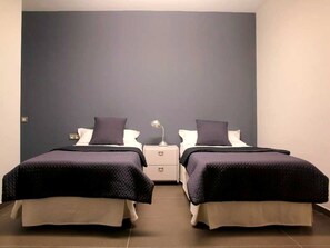 A twin room with two beds, each with a dark-colored bedspread and pillows, a bedside table with a lamp, and a plain wall. - Don Moustache Hostel (Barcelona)