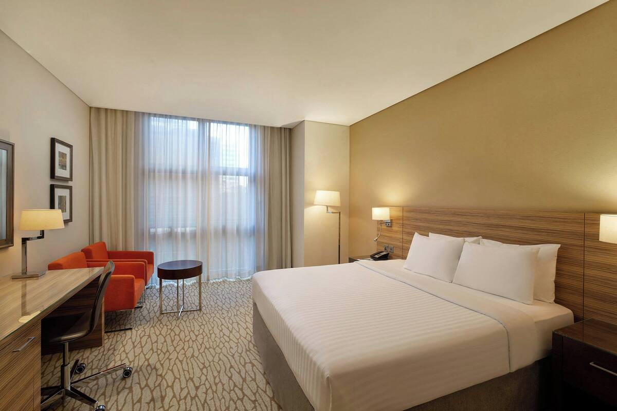 Ein Hotelzimmer mit einem großen Bett, einem Schreibtisch mit Stuhl, einem kleinen Tisch und einem Sofa. An den Wänden befinden sich zwei Lampen, und es gibt ein Fenster mit Vorhängen.