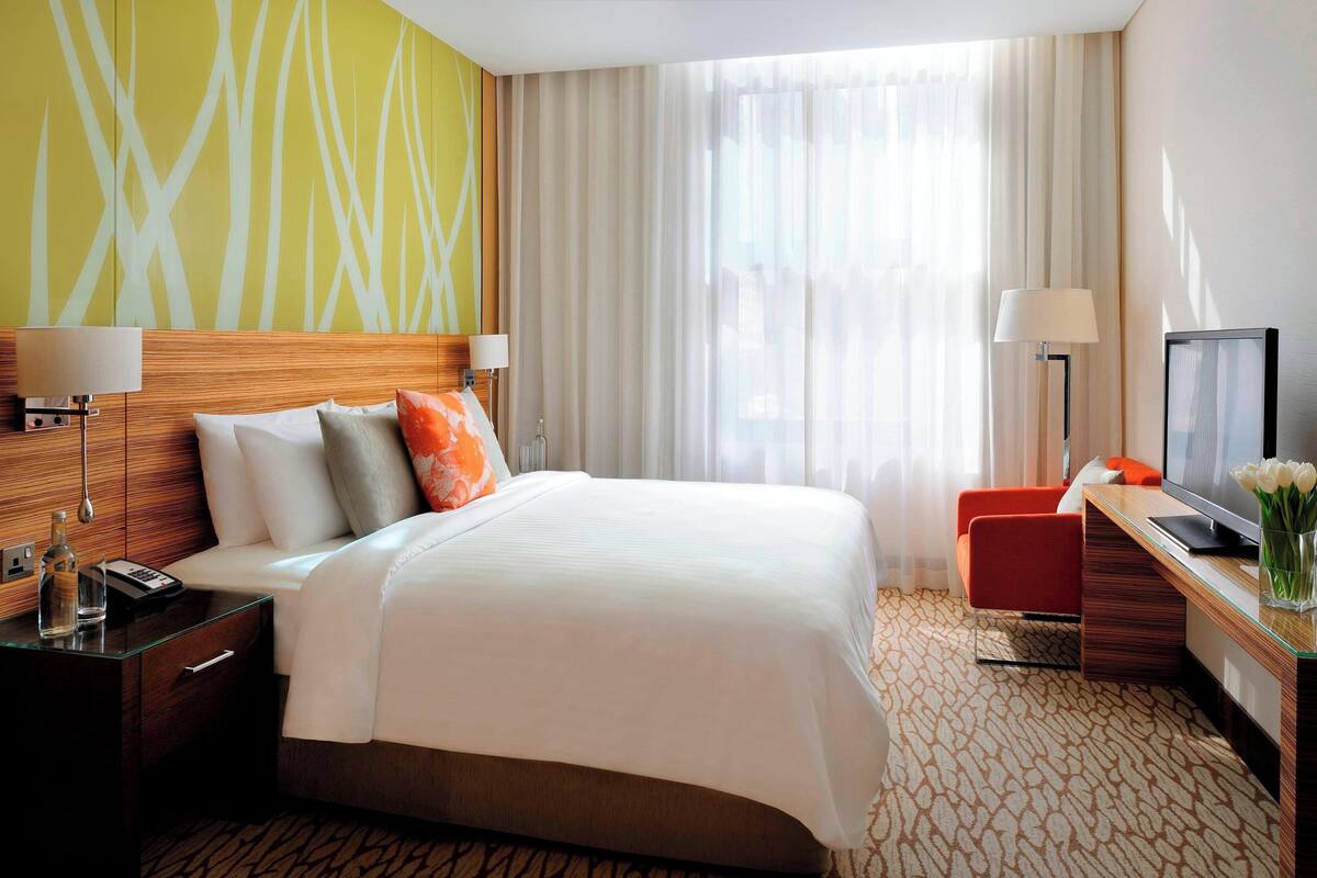 Ein Hotelzimmer mit einem großen Bett, einem Schreibtisch mit Fernseher, einem Sessel und einem Fenster mit Vorhängen.