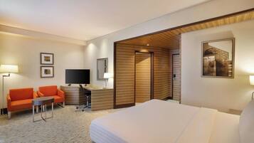 A hotel room with a bed, a sofa, a small table, a desk with a computer, a TV, and a wooden partition wall.