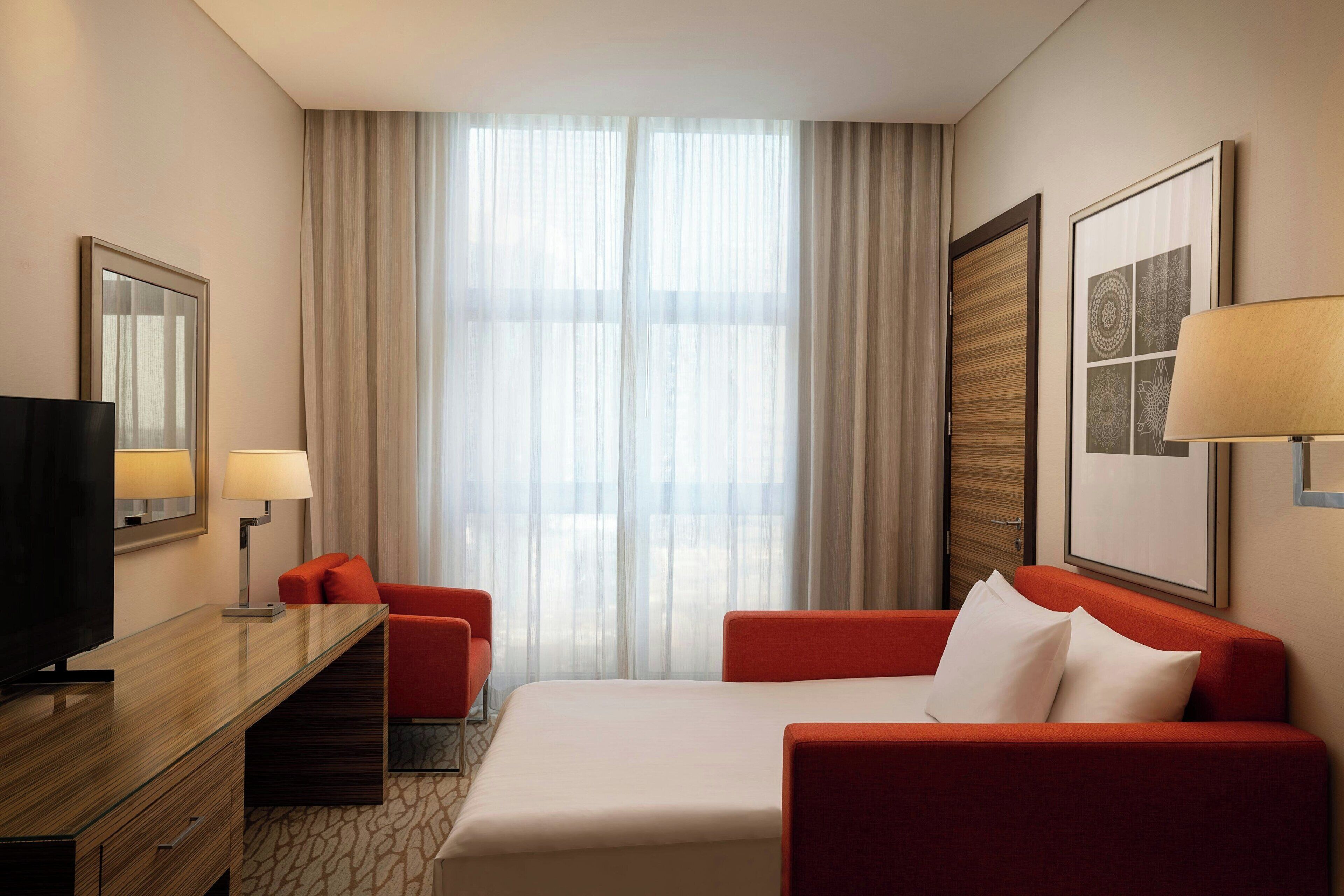 A hotel room with a bed, a desk with a TV, a chair, a lamp, and a large window with curtains.
