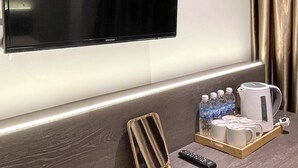 Ein Hotelzimmer mit Flachbildfernseher an der Wand, einem Holztablett mit Wasserflaschen, einer Kaffeemaschine und einer Fernbedienung auf dem Tresen.
