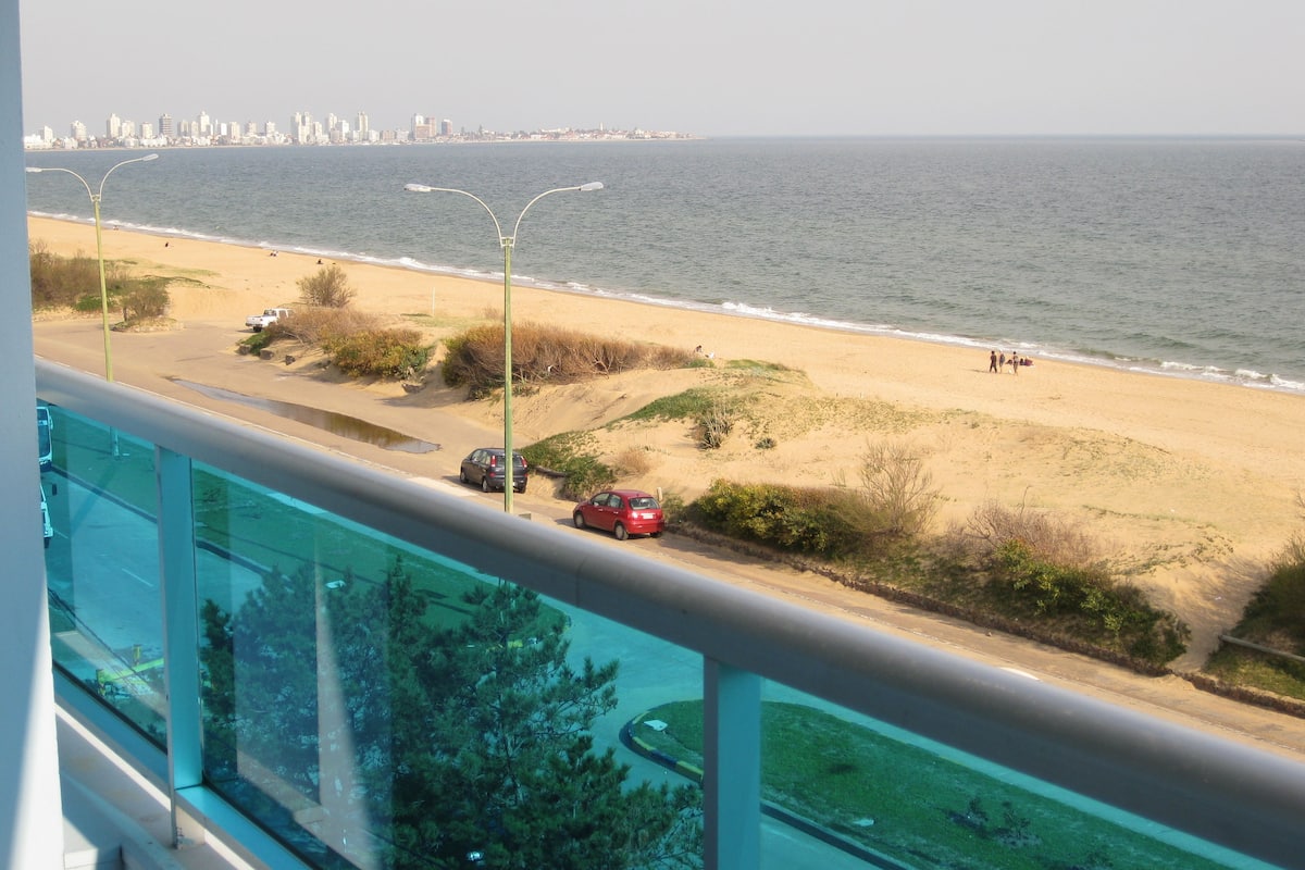 Vista costera desde un balcón con vista a una playa, coches estacionados y el horizonte de la ciudad a lo lejos.