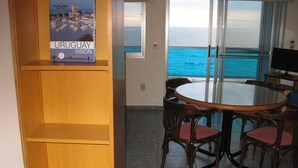 A dining area with a round table and chairs, a book titled "Uruguay Vision" on a shelf, and a view of the ocean through large windows. - Apart Hotel Punta Azul (Punta del Este)