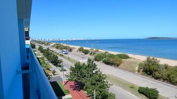 Un paisaje urbano costero con vista despejada a la playa, una carretera con vehículos y un horizonte lejano de la ciudad.