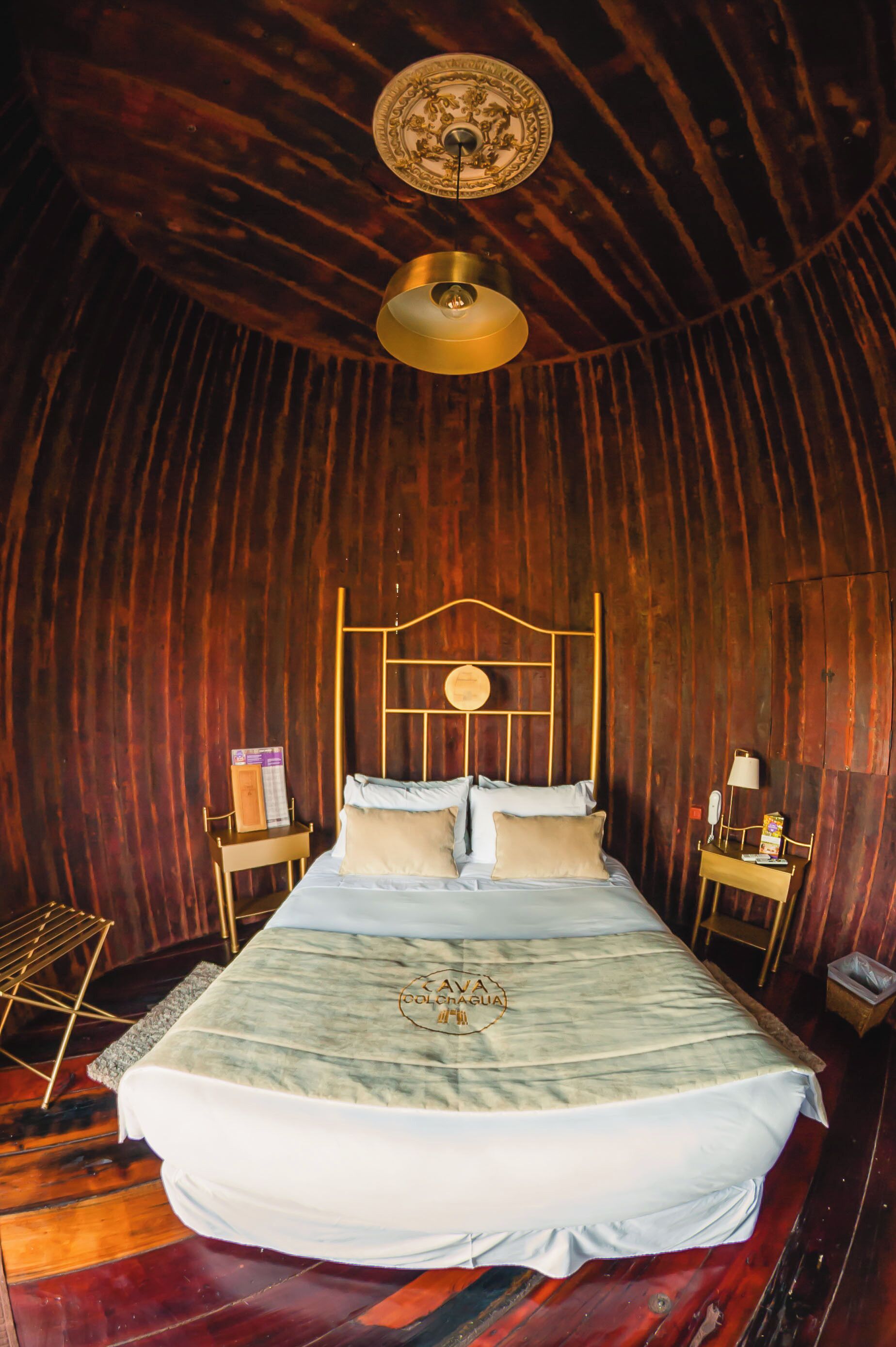 A room with a bed, a wooden ceiling, a small table with a lamp, and a wall-mounted light fixture.