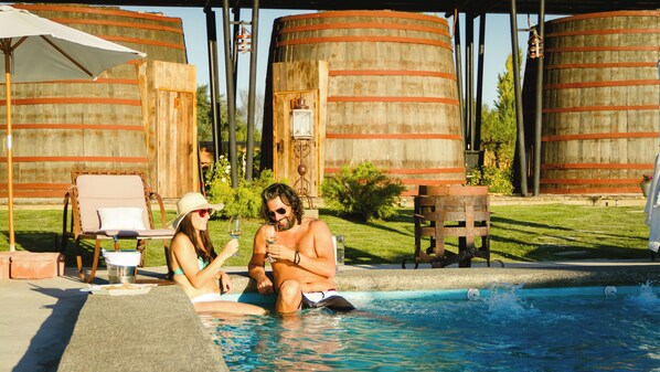 A pool area with two people sitting by the pool, surrounded by wooden barrels and a chair under a white umbrella. - Hotel Cava Colchagua (Santa Cruz)