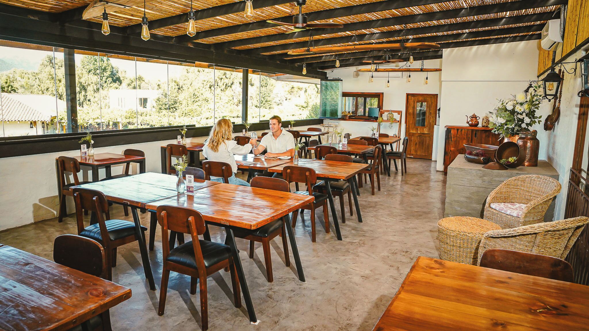 Un comedor amplio con mesas y sillas de madera, ventanas grandes que ofrecen vistas al follaje, y un techo rústico con vigas a la vista y lámparas colgantes.
