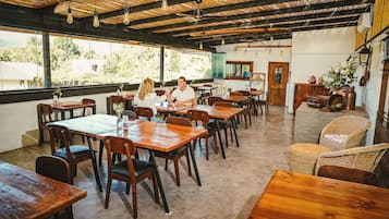 Un comedor amplio con mesas y sillas de madera, ventanas grandes que ofrecen vistas al follaje, y un techo rústico con vigas a la vista y lámparas colgantes.