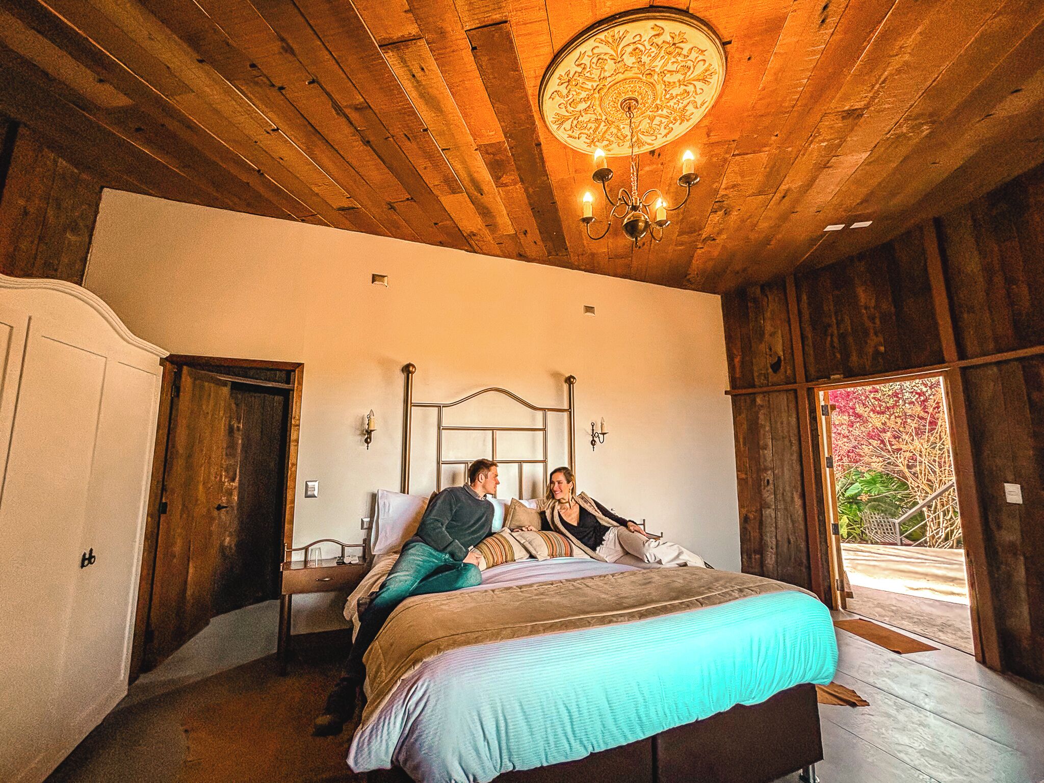 Un dormitorio con techo de madera, un candelabro, una cama con cubrecama azul y vista a zonas verdes al exterior.