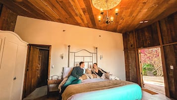 Un dormitorio con techo de madera, un candelabro, una cama con cubrecama azul y vista a zonas verdes al exterior.