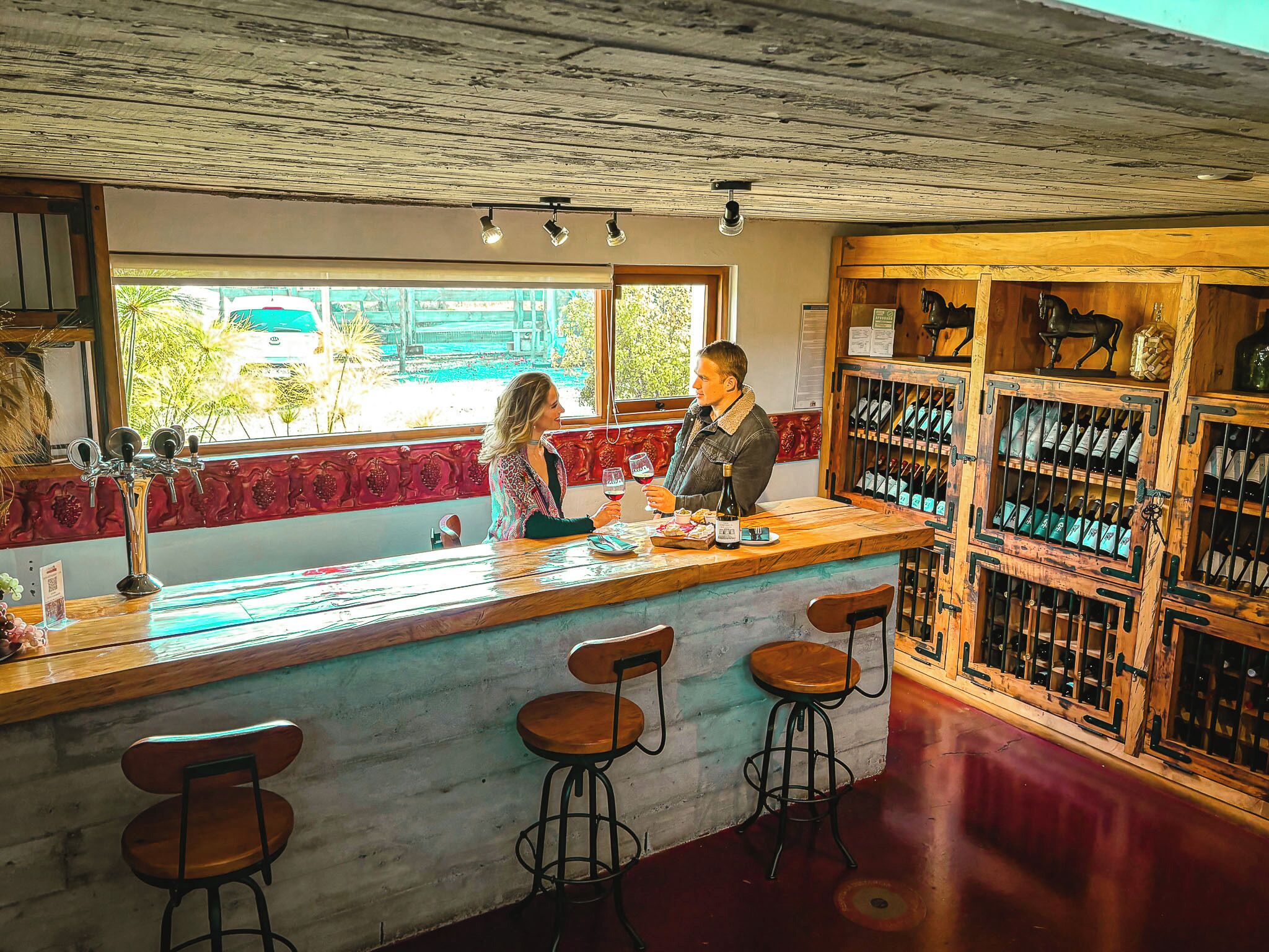 Un bar con mostrador de madera, taburetes y una estantería con botellas de vino.