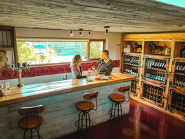 Um bar com balcão de madeira, banquetas e uma estante com garrafas de vinho.
