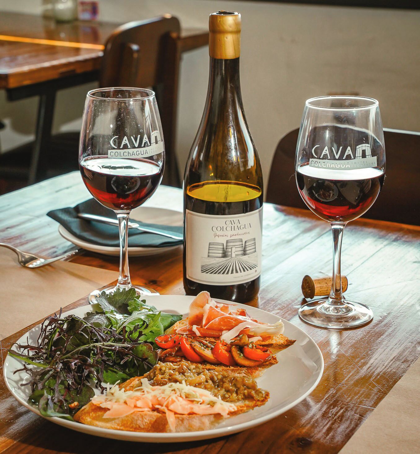 A table with a bottle of wine, two glasses of red wine, and a plate of food.