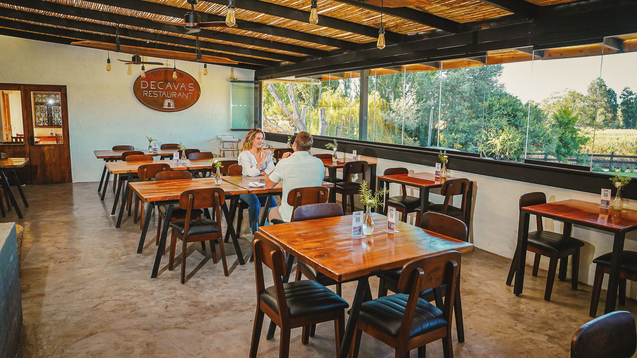 A restaurant interior with wooden tables and chairs, a sign reading 'DECAVAS RESTAURANT', and large windows offering a view of greenery.