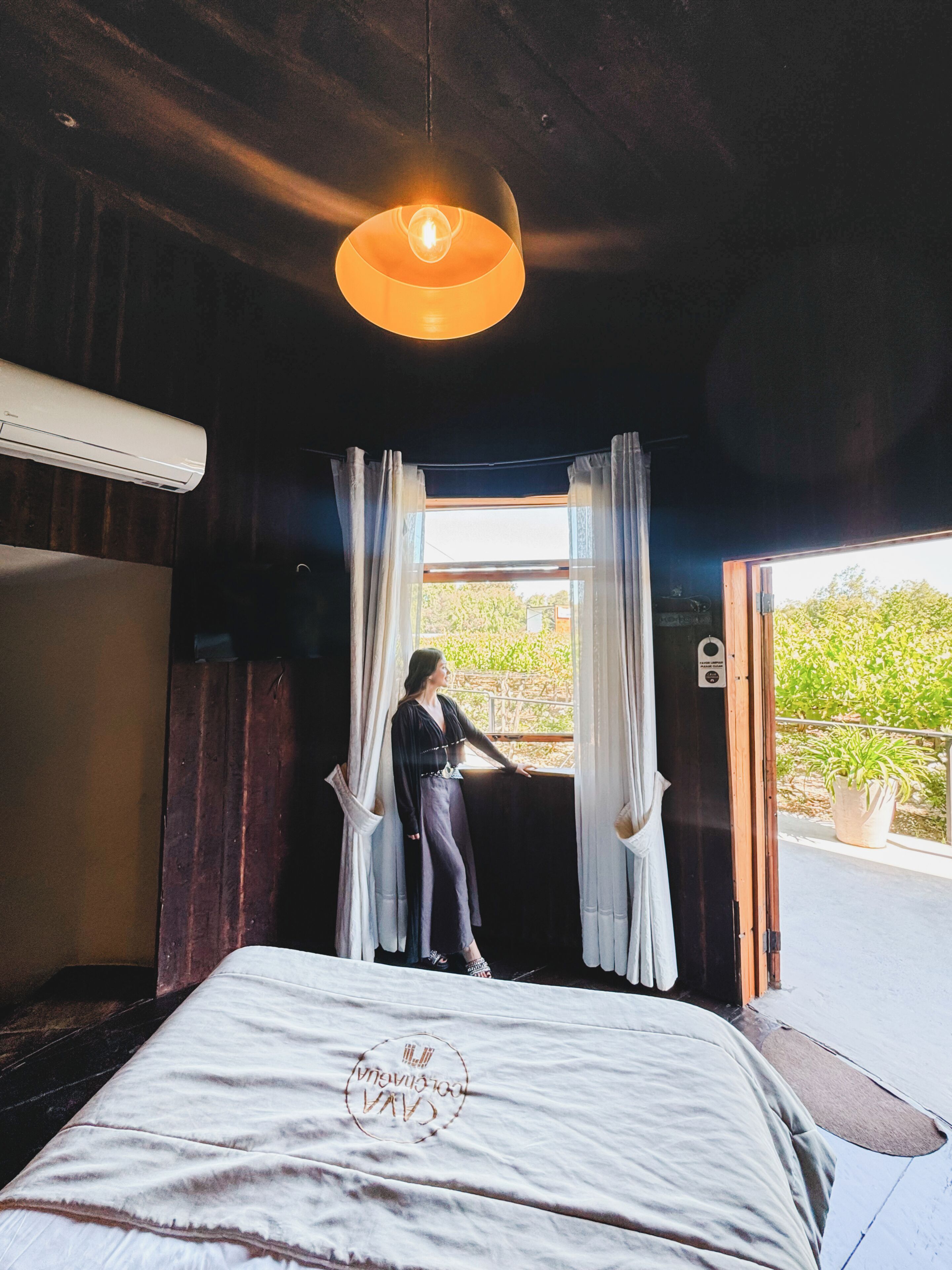 A room with a bed, a window with curtains, a hanging light fixture, and an air conditioning unit.