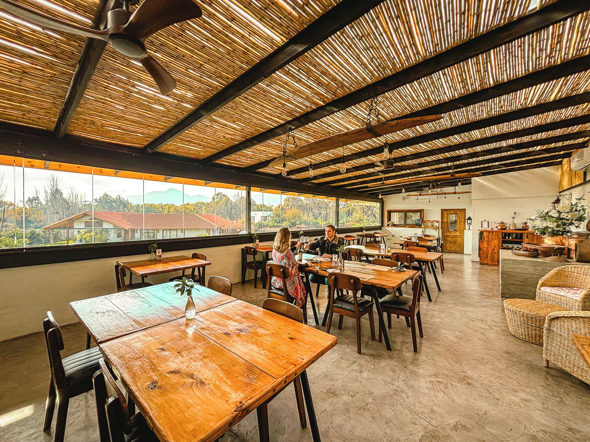 Un comedor interior con mesas y sillas de madera, ventanas amplias y techo de bambú.