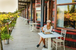 Un área de comedor al aire libre techada, con mesas y sillas blancas, rodeada de plantas en macetas y pilares de madera.