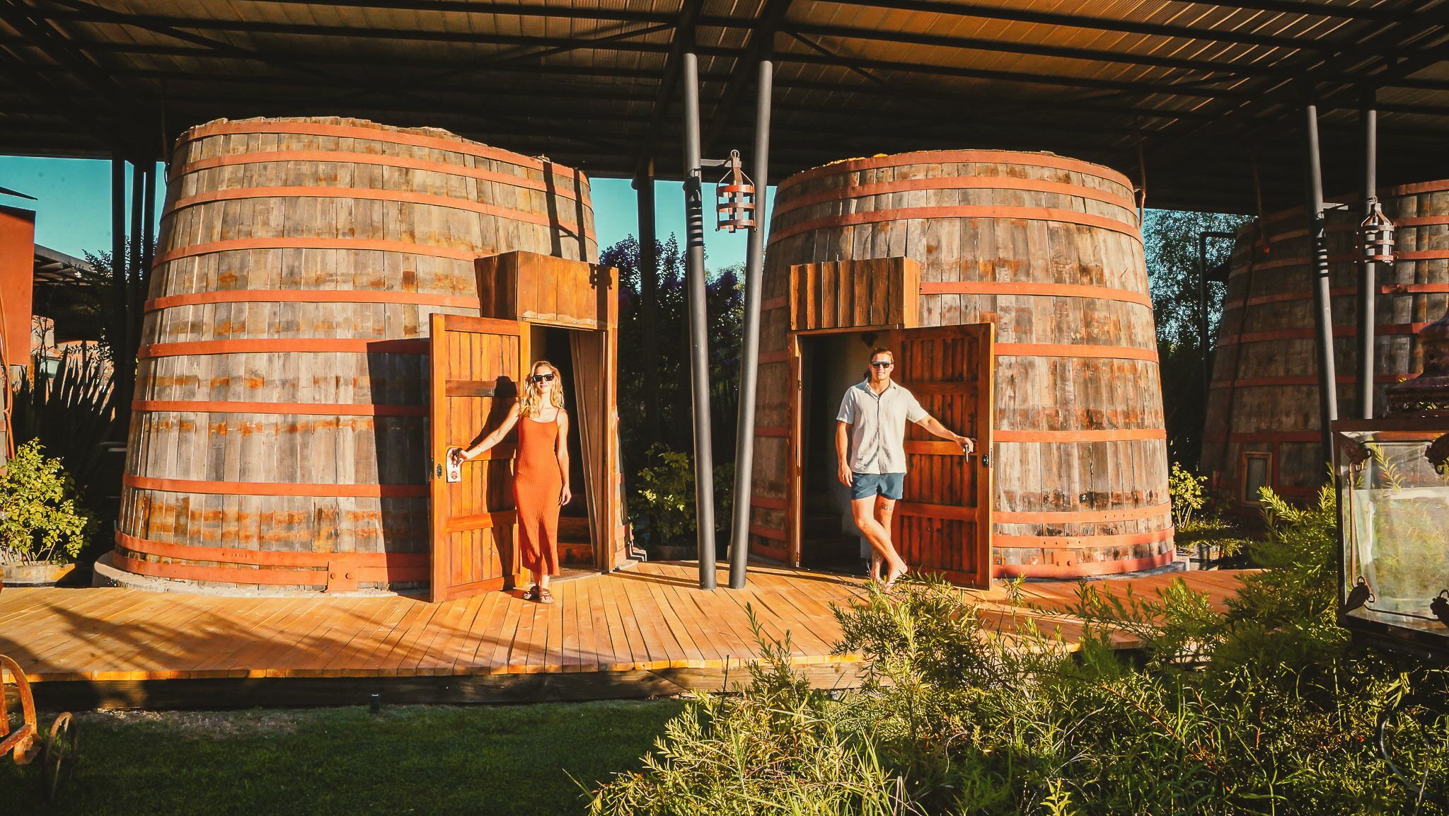 Dos grandes barriles de madera con puertas, un muelle de madera y follaje.