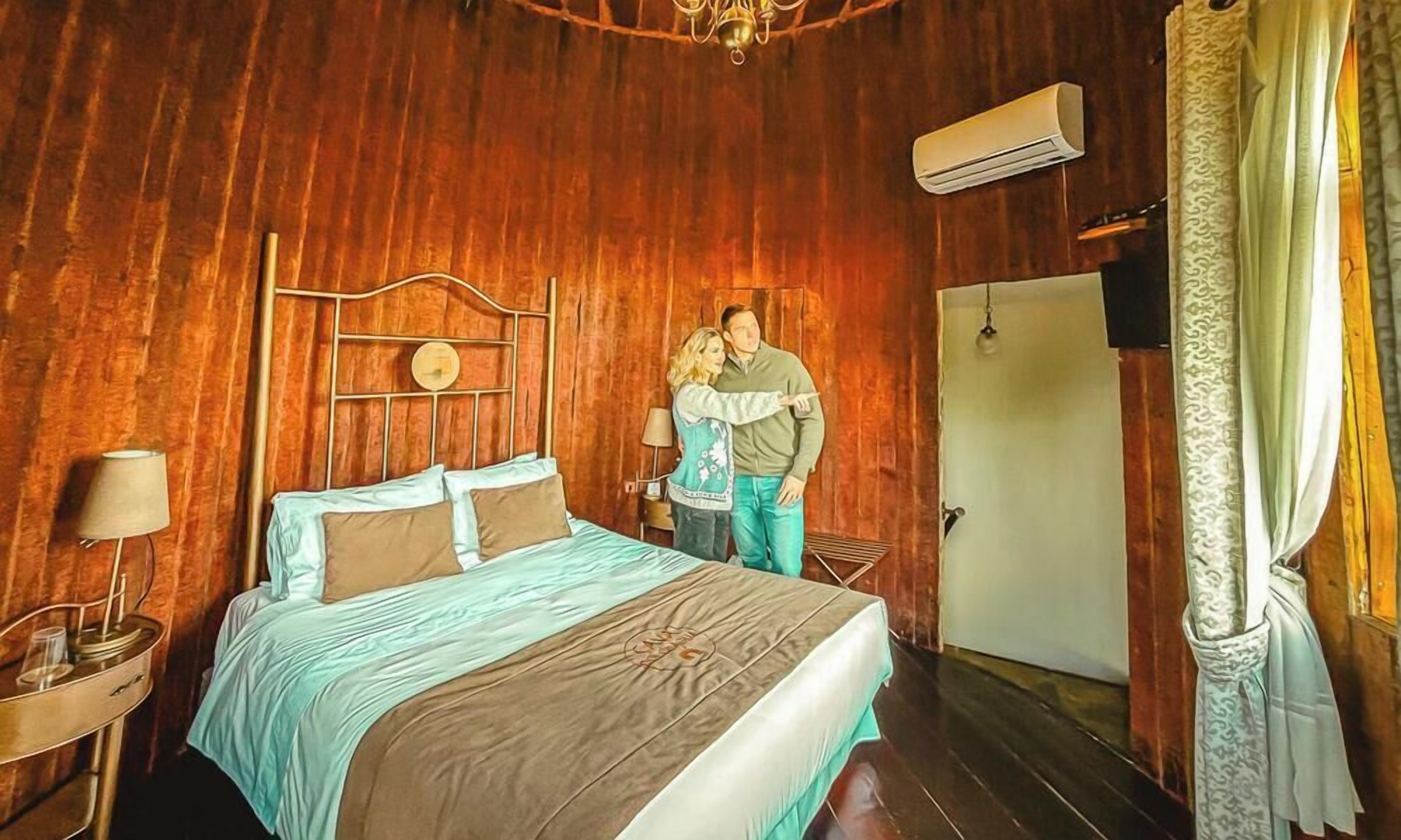 Una habitación con paneles de madera, una cama, una mesita de noche con lámpara y una puerta.