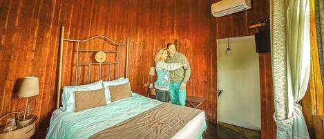 Una habitación con paneles de madera, una cama, una mesita de noche con lámpara y una puerta.