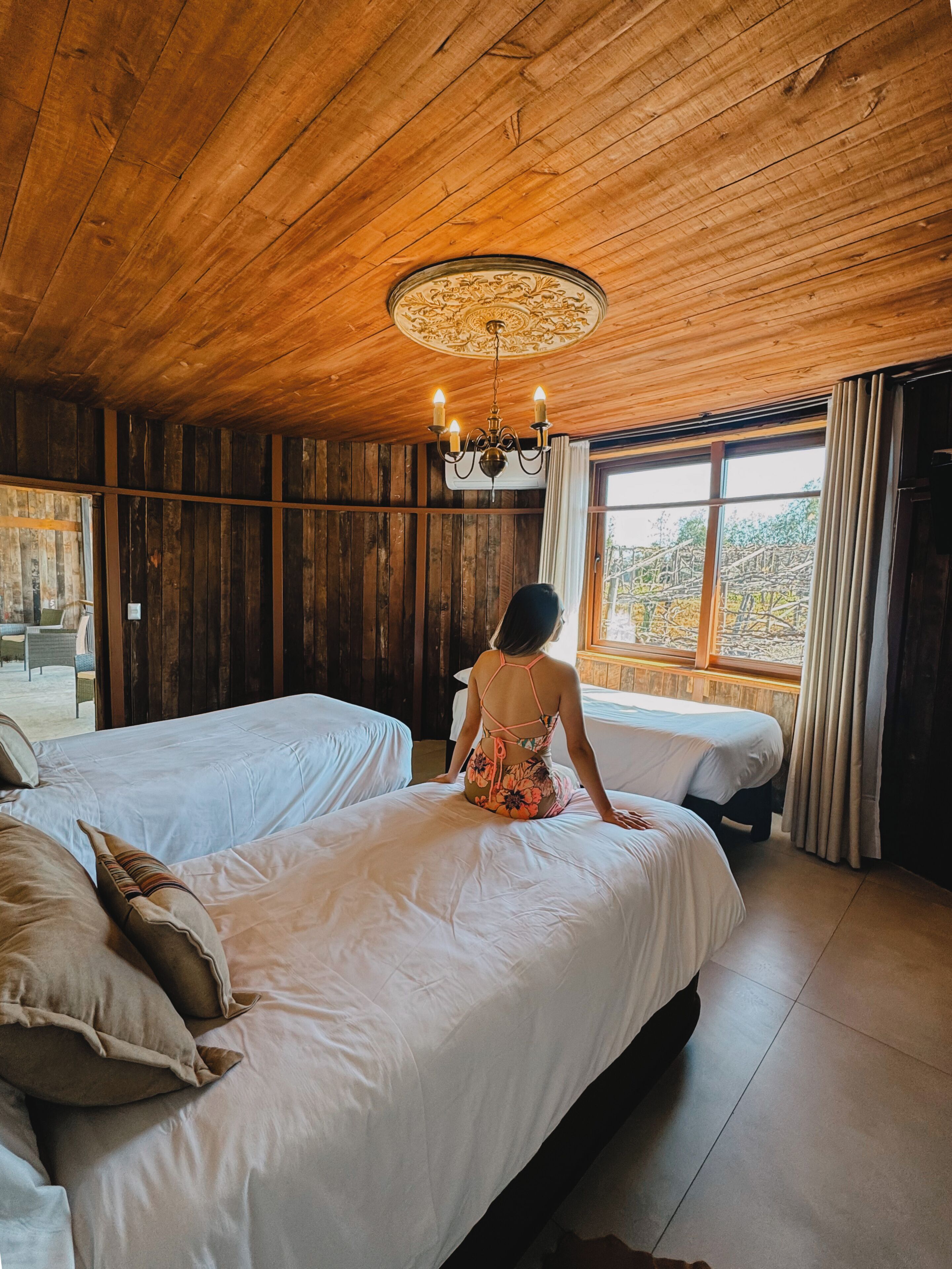 Una habitación con paredes y techo de madera, dos camas, una araña y vistas a través de una ventana.