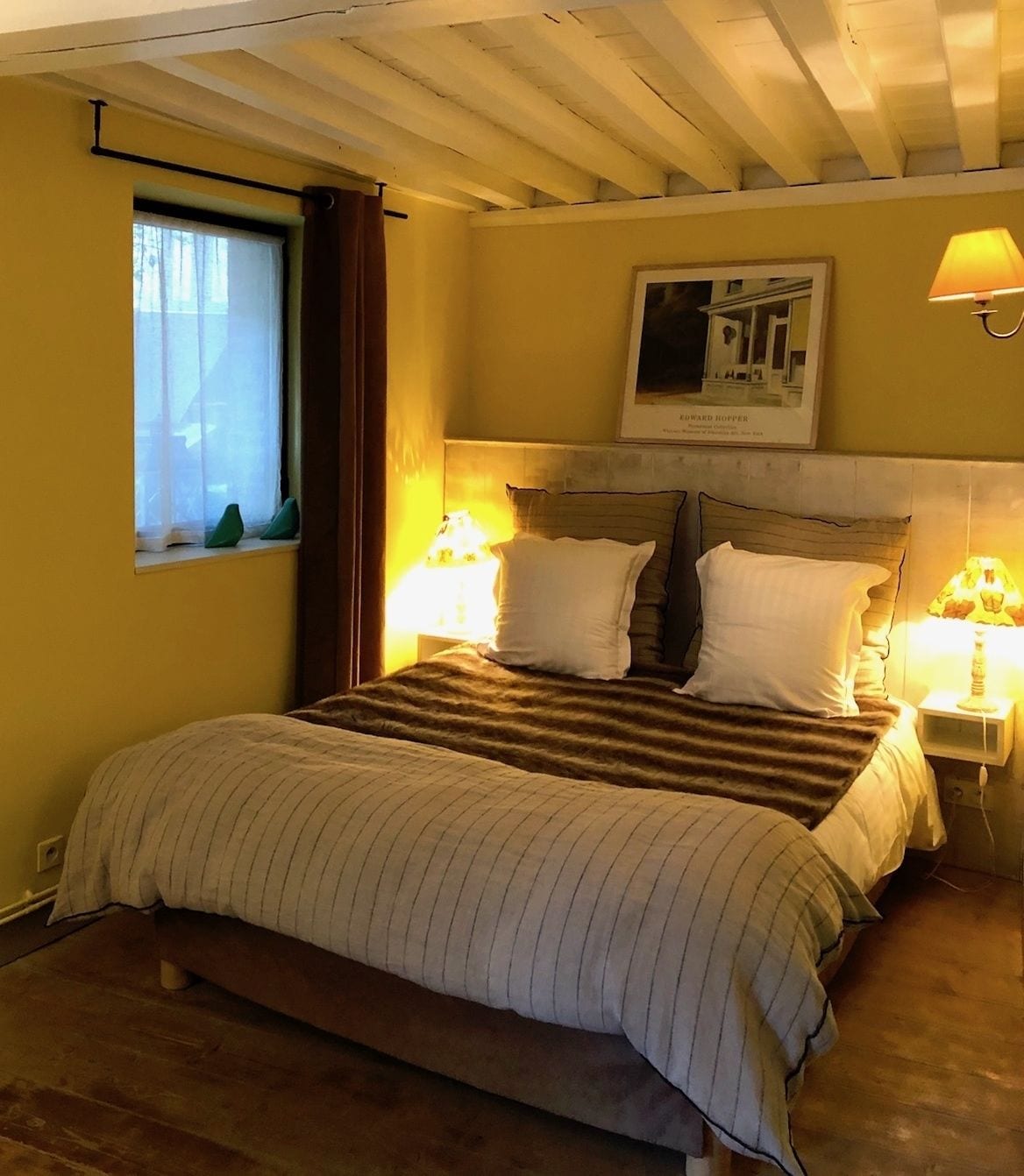 Ein Schlafzimmer mit einem Bett, zwei Nachttischlampen, einem Fenster mit Vorhängen, einem gerahmten Bild an der Wand und einer Holzbalkendecke.