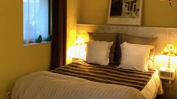 Ein Schlafzimmer mit einem Bett, zwei Nachttischlampen, einem Fenster mit Vorhängen, einem gerahmten Bild an der Wand und einer Holzbalkendecke.