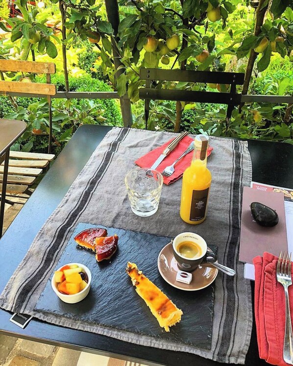 Mesa posta ao ar livre com uma refeição que inclui uma cesta de frutas, um pastel, um copo de água, uma garrafa de suco, uma xícara de café e um guardanapo.