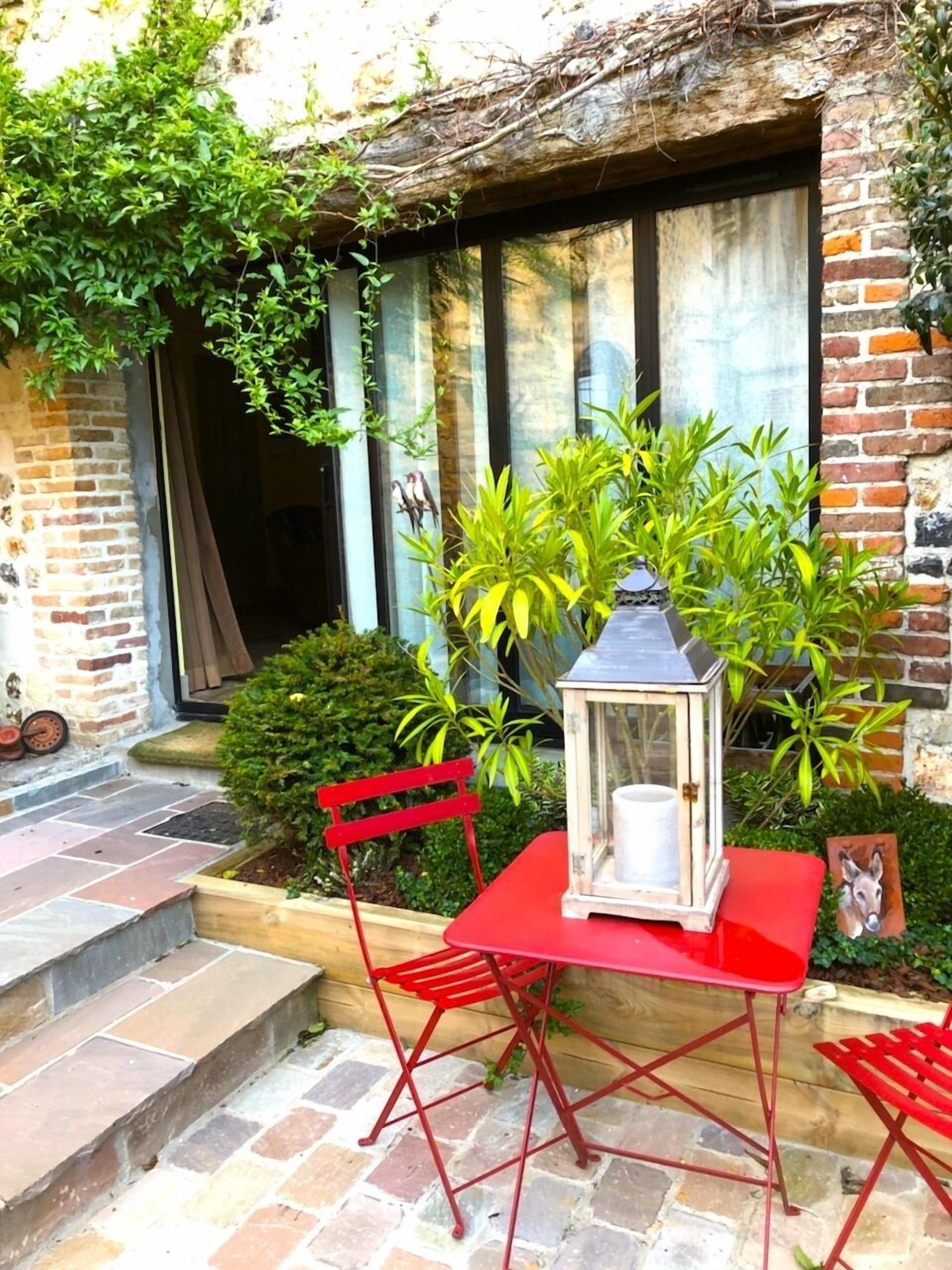 Un conjunto de mesa y sillas rojas situado afuera de un edificio de ladrillo con una ventana y un pequeño jardín.