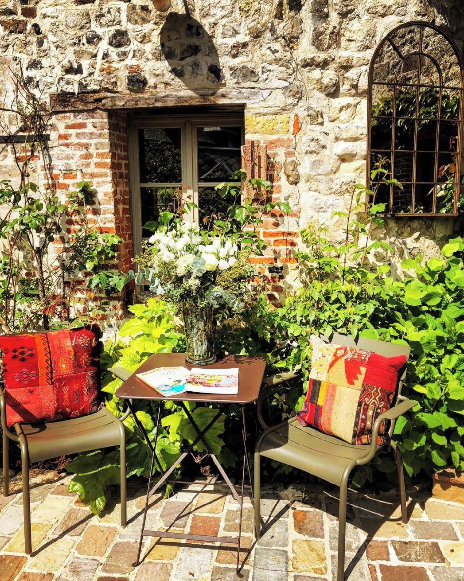 Un edificio de piedra con una ventana y una puerta, dos sillas de metal con cojines, una mesa con un libro y un jarrón con flores.