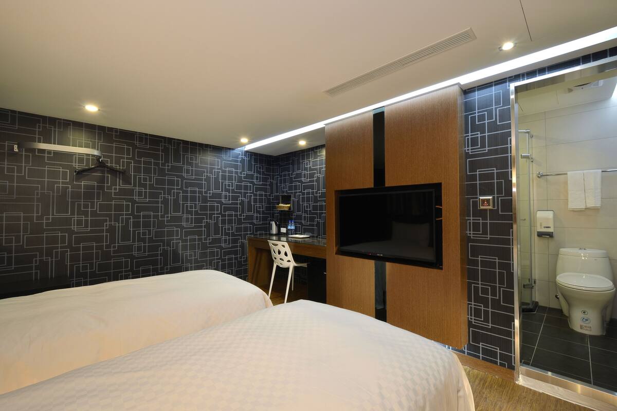 A hotel room with two beds, a desk with a chair, a TV, and a shower area.