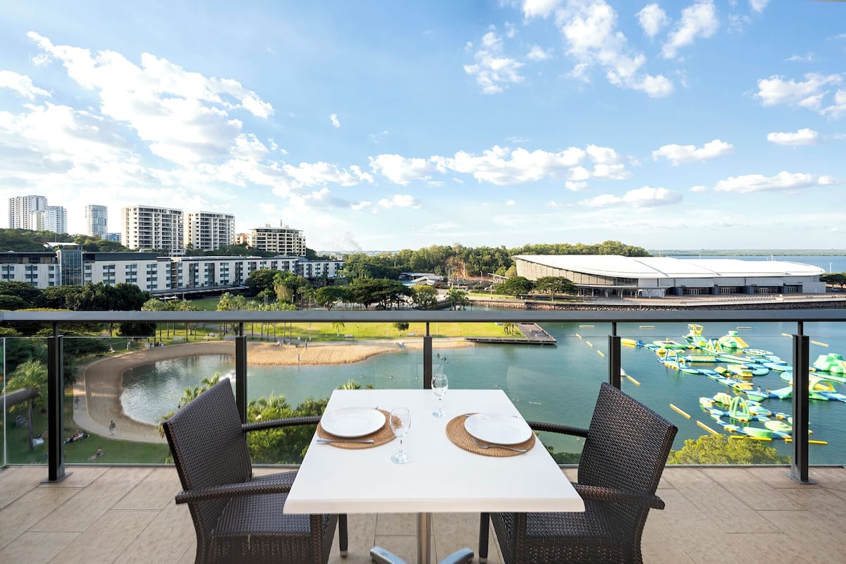 Un balcón con una mesa para dos, con vista a un cuerpo de agua con barcos y un paisaje urbano de fondo.