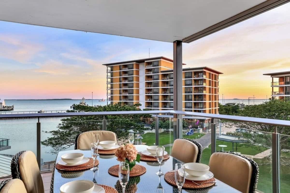 Comedor con una mesa dispuesta para dos personas, con vistas a una masa de agua y un conjunto de edificios.