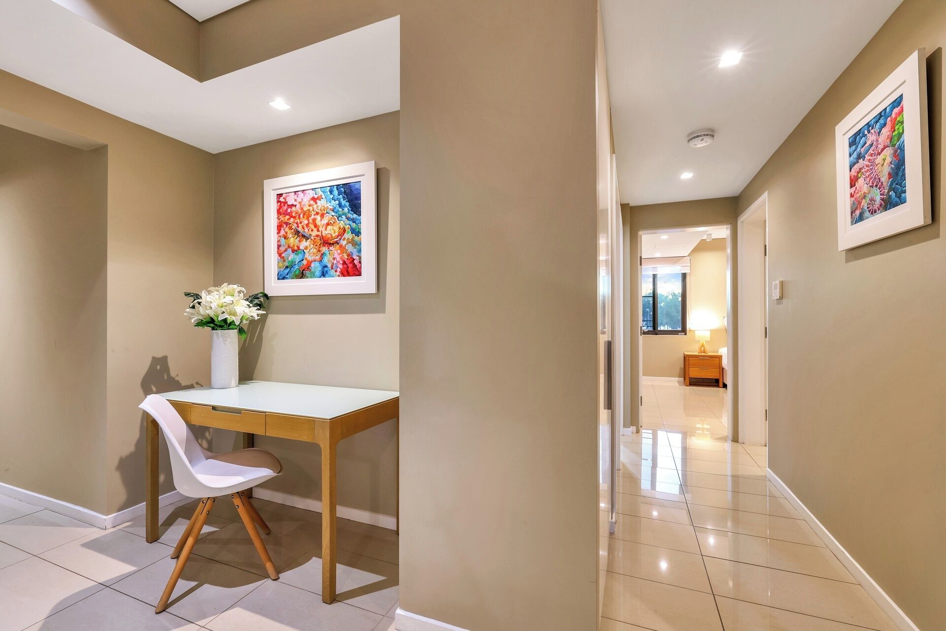 A hallway with a desk and chair, a vase of flowers, and two framed artworks on the walls.