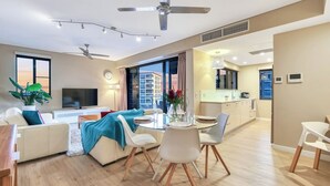 Een moderne woonkamer met een bank, televisie, eettafel en stoelen. Er is een keuken met kasten en een raam.