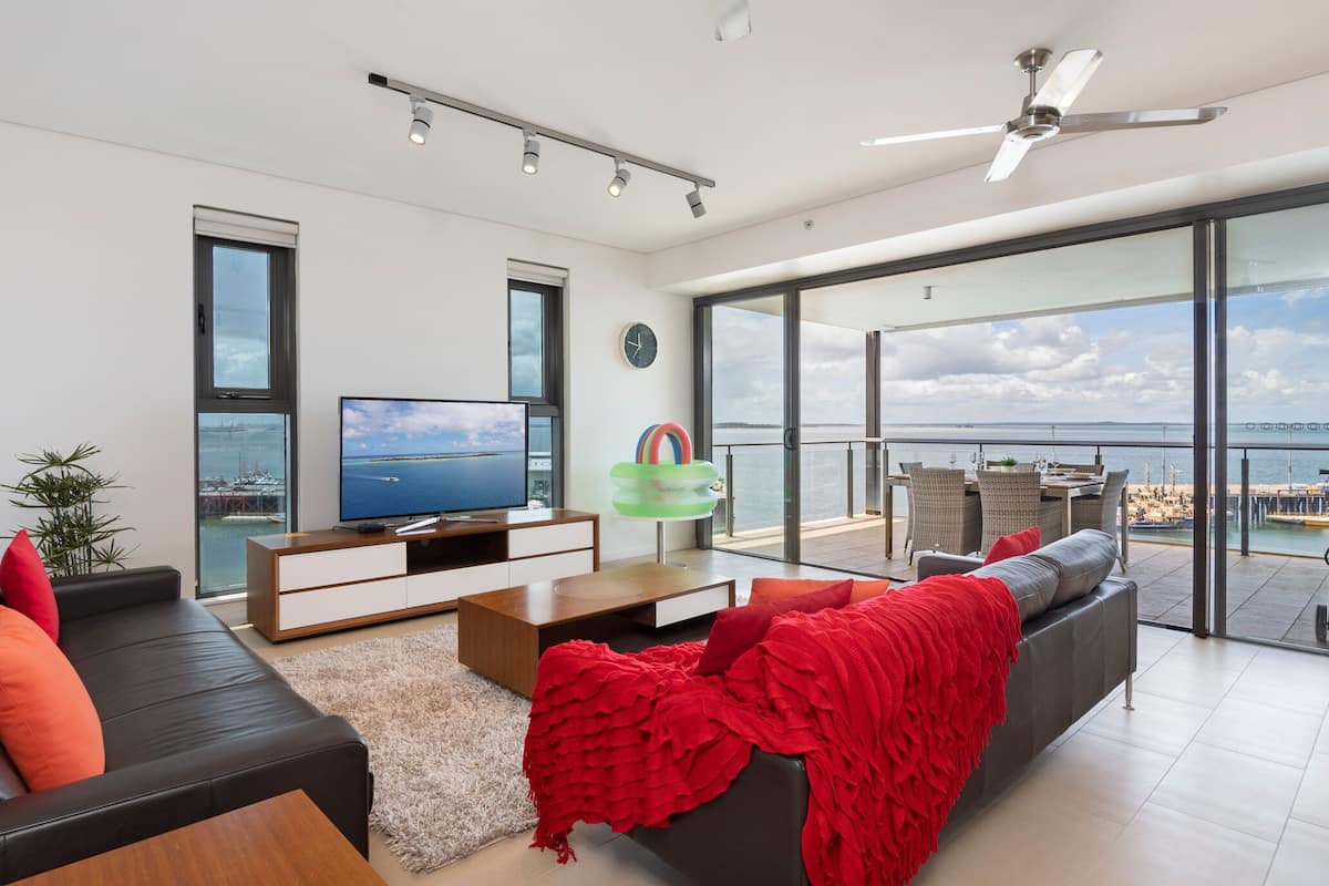 Una sala de estar moderna con un televisor grande, asientos cómodos, un balcón con vista al mar y un ventilador de techo.