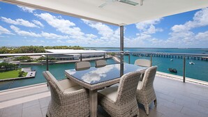 Een balkon met een glazen tafel en rieten stoelen, met uitzicht op een jachthaven met boten en een steiger.