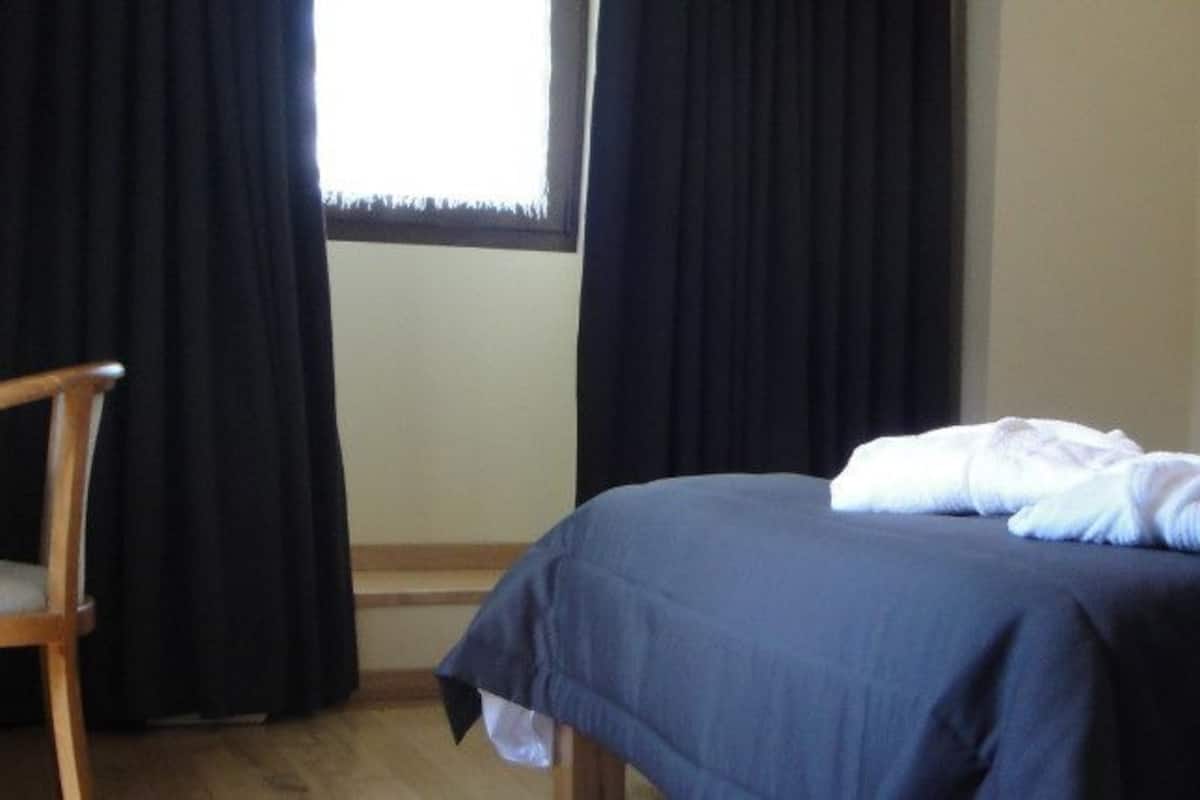 Una habitación de hotel con cama, una silla, una ventana con cortinas y una lámpara fijada a la pared.