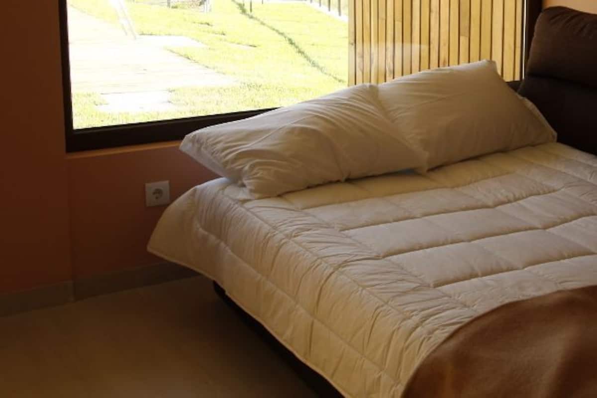 Una cama con edredón blanco y almohadas, una manta marrón y una ventana con persianas.