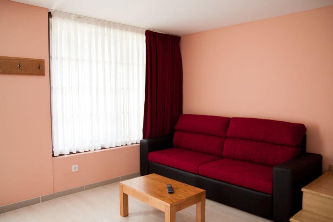 A living room with a red sofa, a wooden coffee table, and a window with sheer curtains and a red drape.