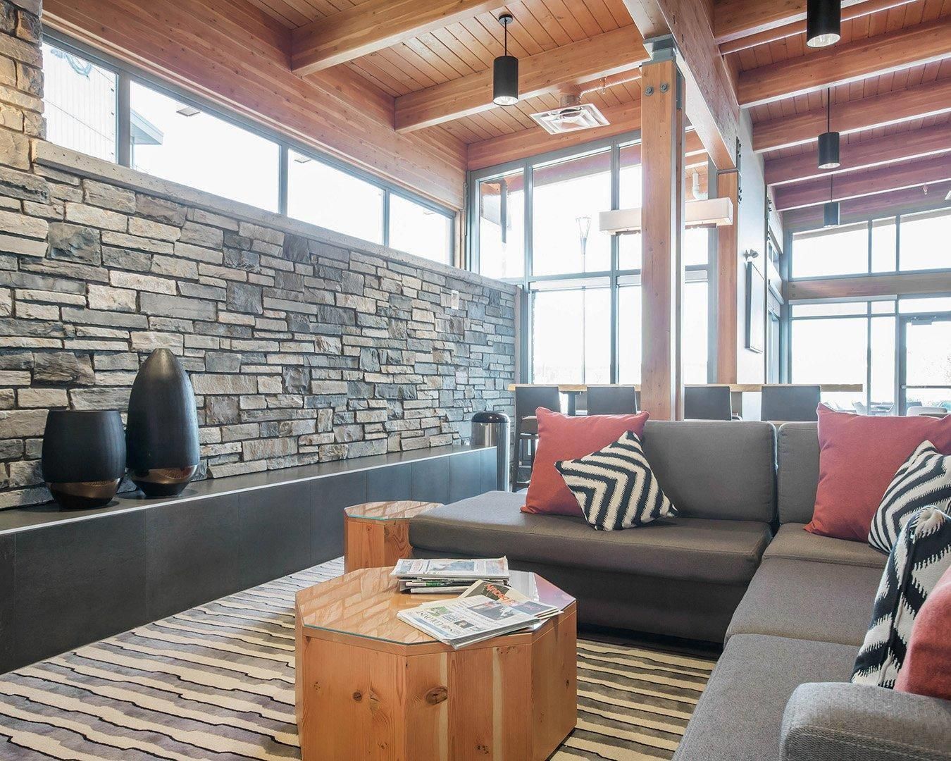 A modern interior with a stone wall, wooden ceiling beams, a sofa with patterned cushions, and a coffee table with magazines.