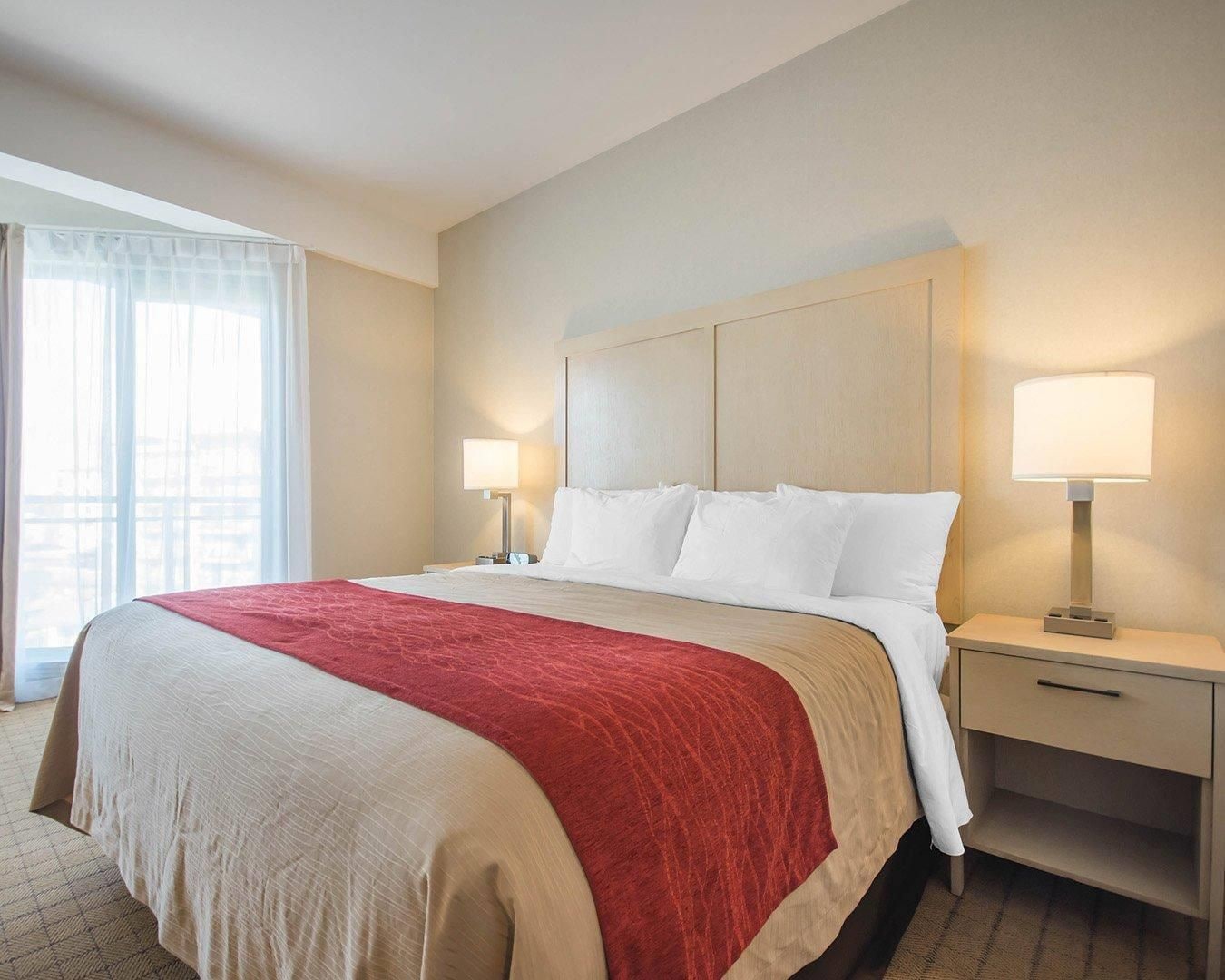 A hotel room with a large bed, two bedside tables with lamps, a nightstand, and a view of the city through large windows.