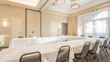 Un salón de conferencias con mesas y sillas largas, un amplio ventanal con cortinas y un techo con luminarias circulares.