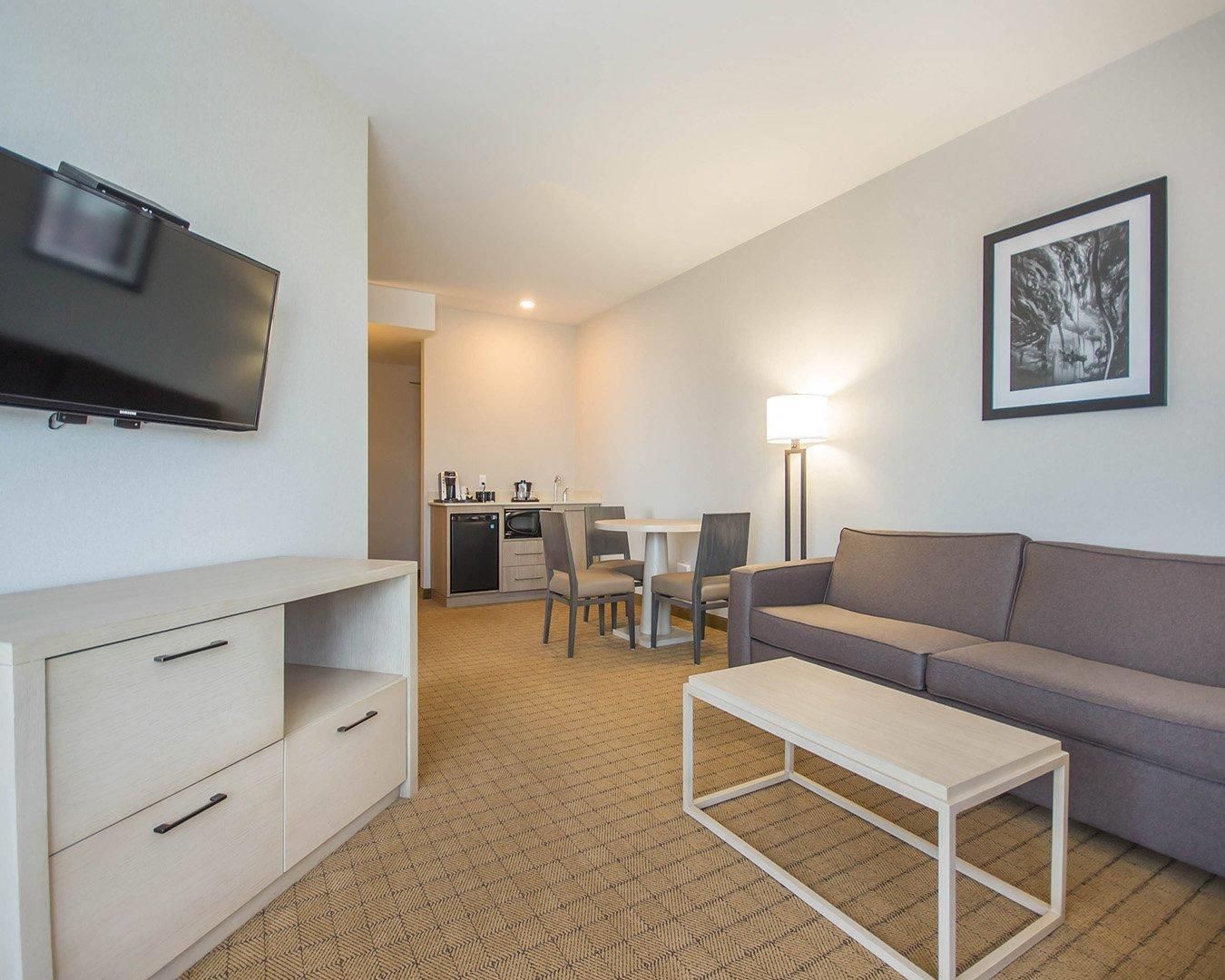 A hotel room with a flat-screen TV, a sofa, a dining table with chairs, a coffee machine, and a lamp.