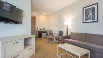 A hotel room with a flat-screen TV, a sofa, a dining table with chairs, a coffee machine, and a lamp.