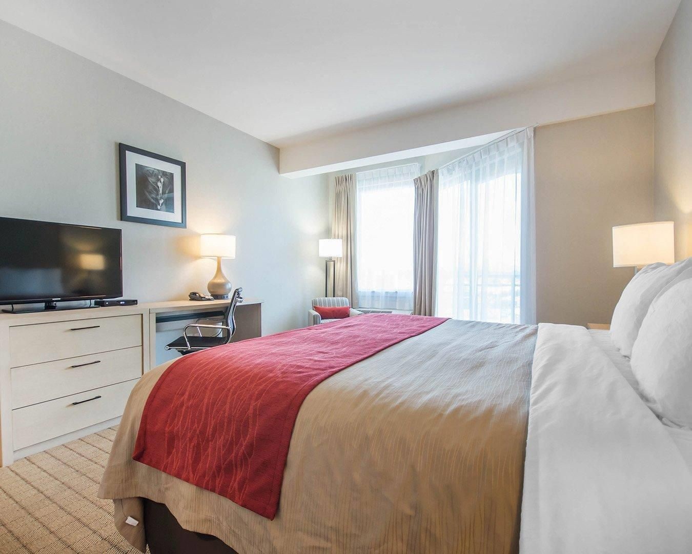 A hotel room with a bed, a desk with a TV, a chair, and a window with curtains.