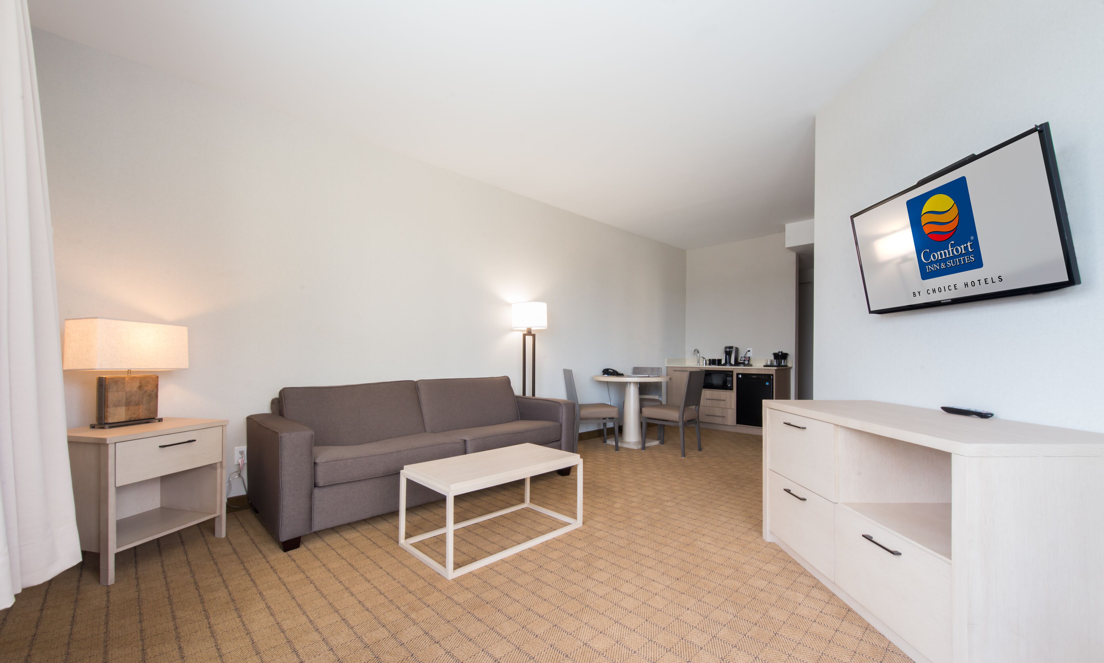 A compact hotel room with a grey sofa, a small white table, a desk with a chair, a kitchenette, and a framed Comfort Inn & Suites sign.