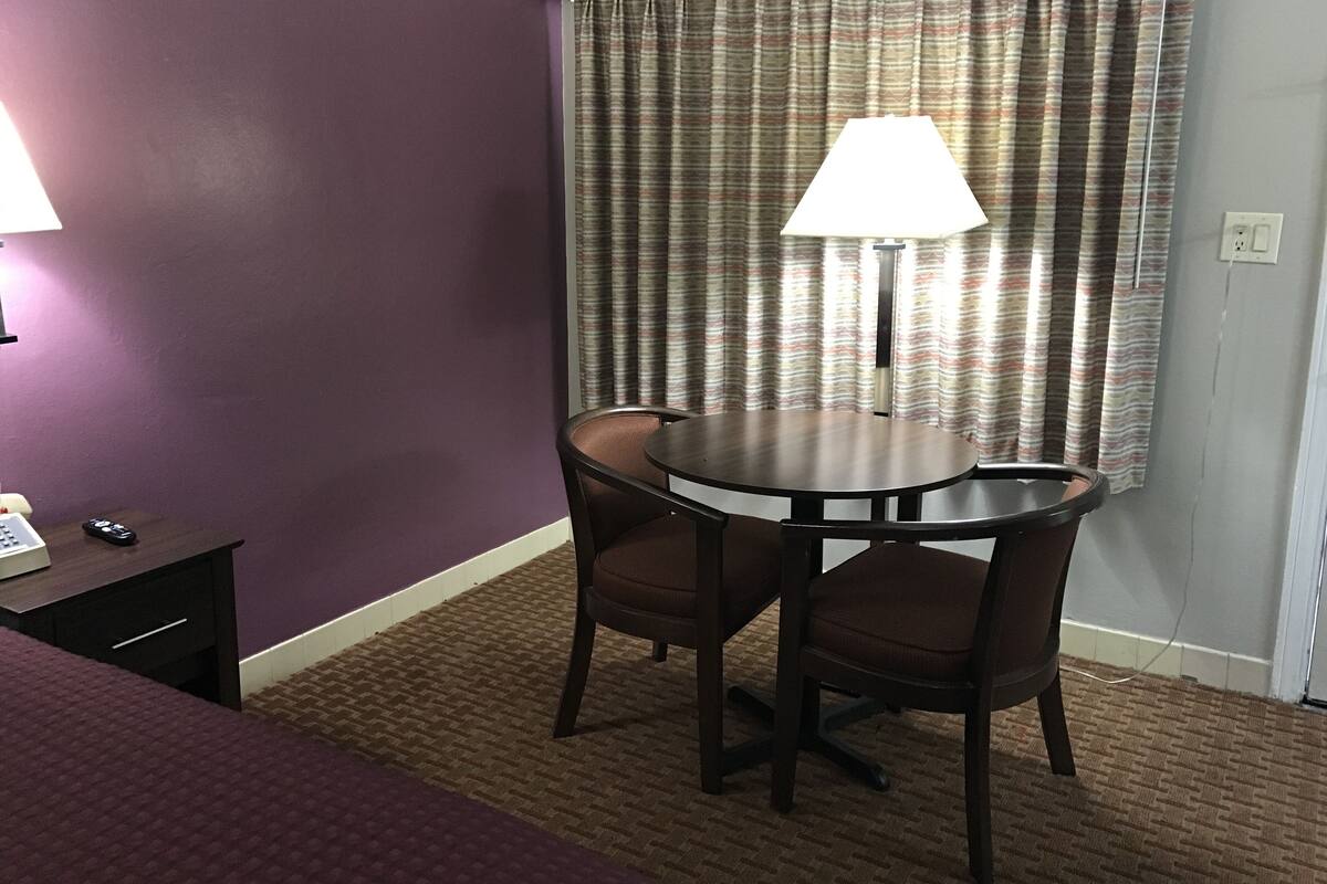A hotel room with a round table, two chairs, a lamp, a small desk with a phone, and a purple wall.