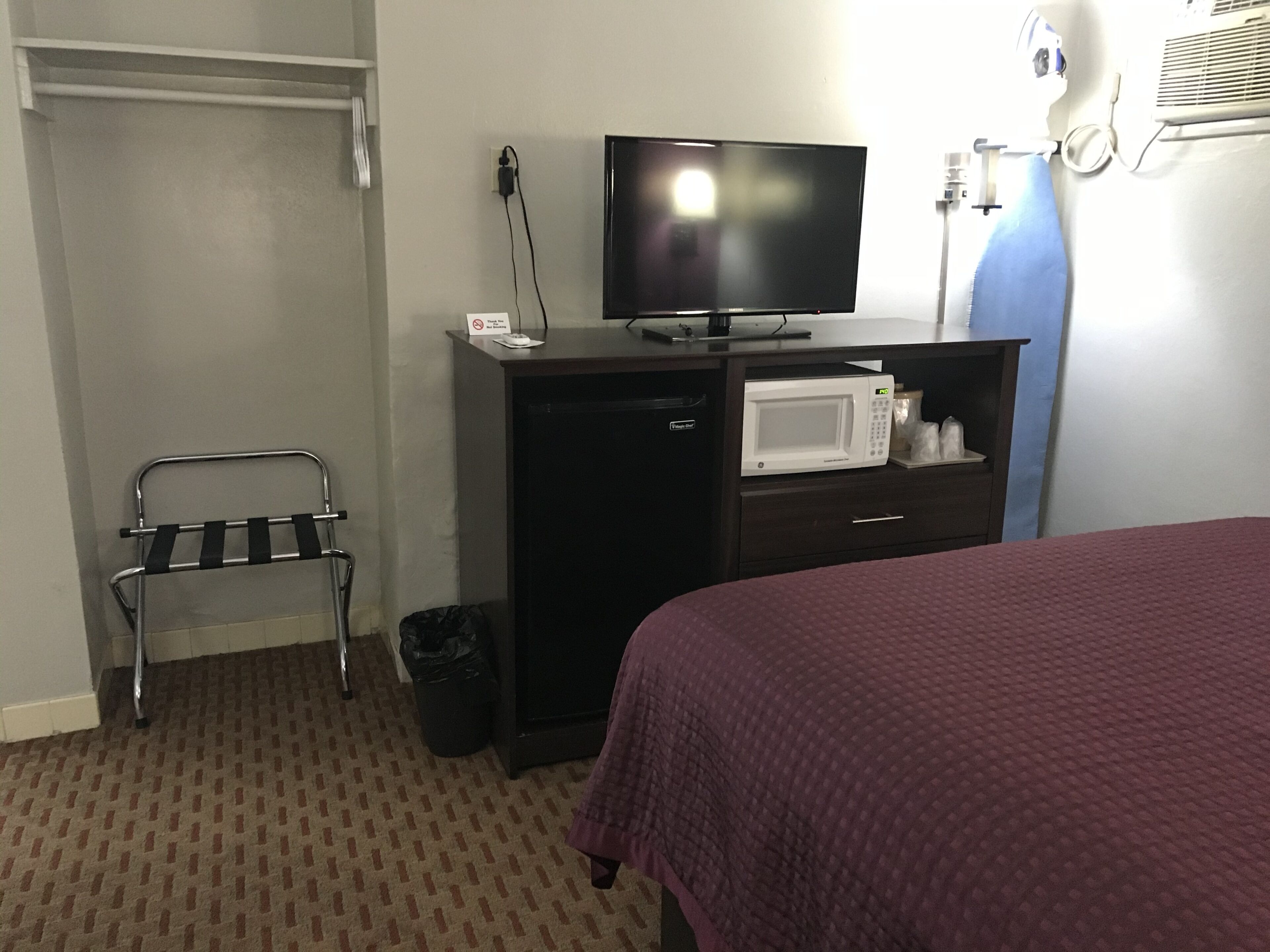 A hotel room with a bed, a television, a microwave, a small desk, a trash can, and a shoe rack.